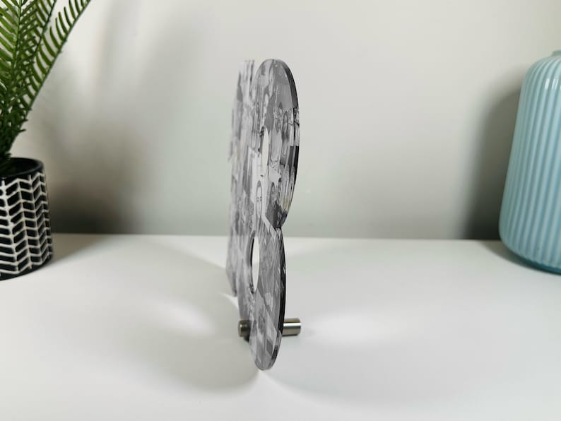 K&ouml;nnte beinhalten: Ein Schwarzwei&szlig;-Fotocollage in Form des Buchstabens "B" ist auf einem klaren Acryl-St&auml;nder montiert. Der St&auml;nder hat eine silberne Metallbasis.