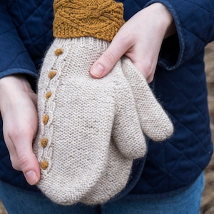 Puede incluir: Un par de mitones de punto beige con un puño de punto de cable marrón y botones marrones en el lateral.