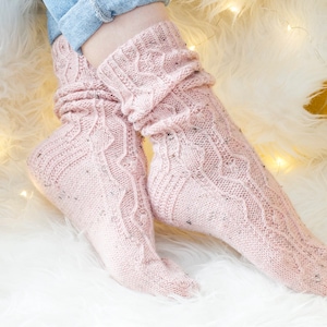 May include: Pale pink knitted socks with a cable knit design and small, sparkly details. The socks are worn, with the legs slightly scrunched. The background features a white fluffy rug and a yellow surface.