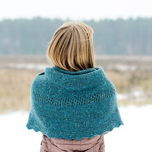 Puede incluir: Una mujer con un chal de punto azul turquesa con un patrón de encaje se encuentra en un campo nevado con un fondo borroso de árboles.