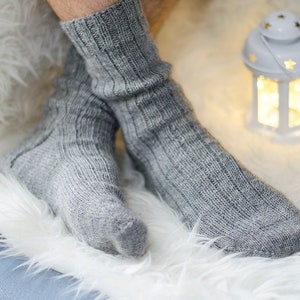 Pode incluir: Meias cinzentas de malha com um design canelado. As meias estão sobre uma superfície branca e fofa. Uma pequena lanterna branca com recortes de estrelas está no fundo.