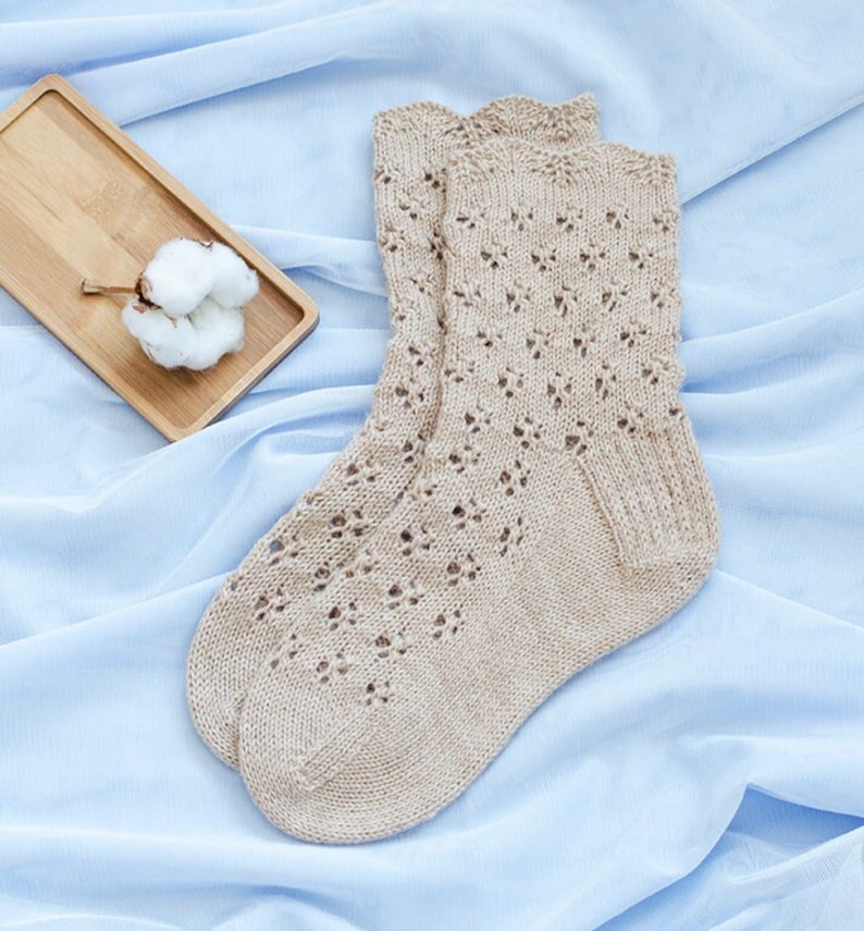 May include: A pair of beige knitted socks with a delicate lace pattern. The socks are laid out on a light blue fabric background.