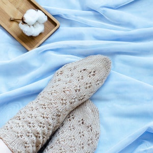 May include: A pair of beige knitted socks with a lacy pattern, resting on a light blue fabric background. A small wooden tray with a cotton boll sits in the upper left corner of the image.