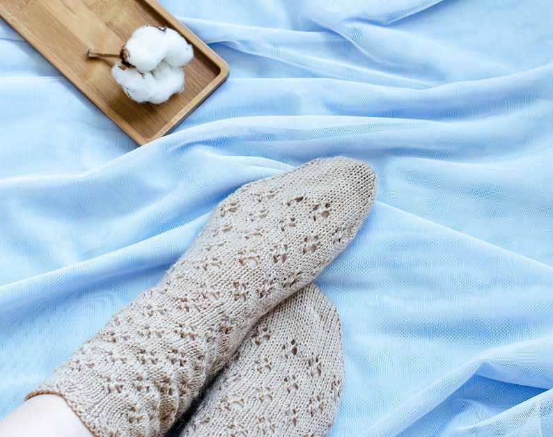 May include: A pair of beige knitted socks with a lacy pattern, resting on a light blue fabric background. A small wooden tray with a cotton boll sits in the upper left corner of the image.