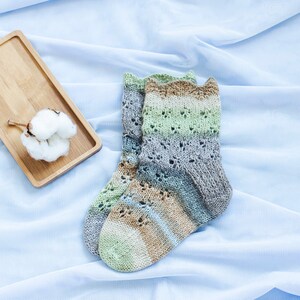 May include: A pair of knitted socks with a brown, green, and gray color pattern. The socks have a lacy design and are lying on a light blue fabric background.