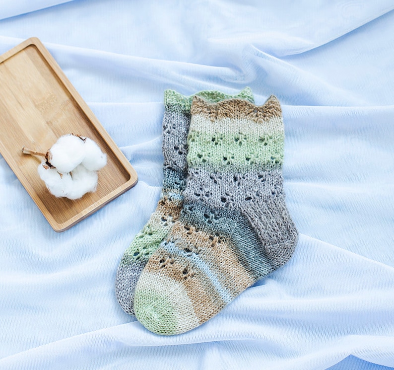 May include: A pair of knitted socks with a brown, green, and gray color pattern. The socks have a lacy design and are lying on a light blue fabric background.