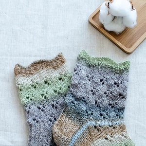May include: A pair of hand-knitted socks in a green, brown, and gray color scheme with a lacy pattern. The socks are on a white linen background.