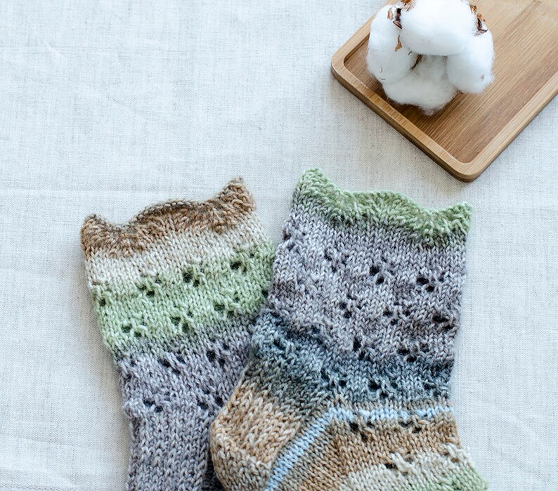 May include: A pair of hand-knitted socks in a green, brown, and gray color scheme with a lacy pattern. The socks are on a white linen background.