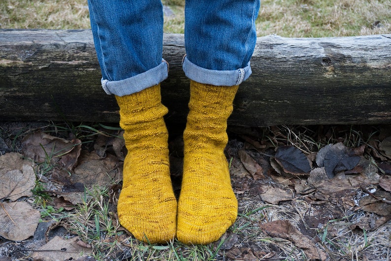 K&ouml;nnte beinhalten: Ein Paar gelbe Stricksocken mit einem strukturierten Muster. Die Socken werden von einer Person getragen, die eine blaue Jeans tr&auml;gt.