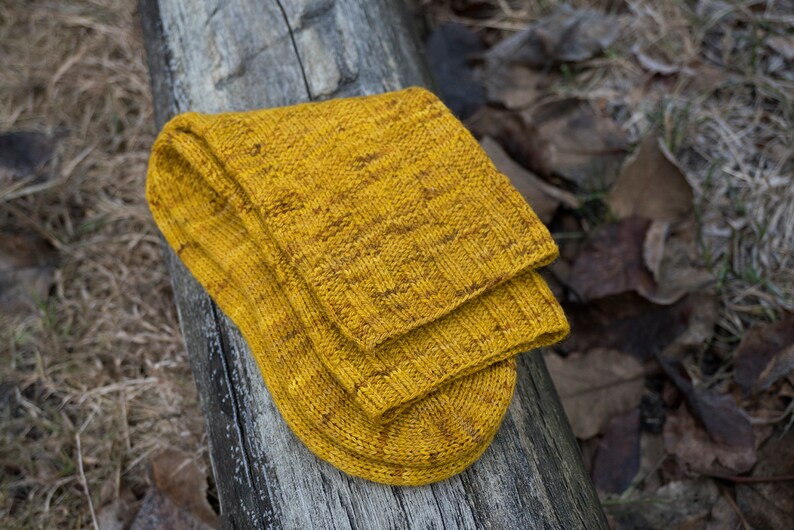 K&ouml;nnte beinhalten: Ein Paar gelbe Stricksocken mit einem strukturierten Muster. Die Socken sind gefaltet und liegen auf einer Holzoberfl&auml;che.