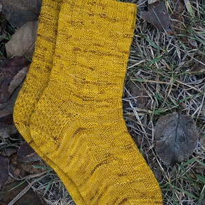 K&ouml;nnte beinhalten: Ein Paar gelbe Stricksocken mit einem strukturierten Muster. Die Socken liegen auf einem Bett aus trockenen Bl&auml;ttern und Gras.