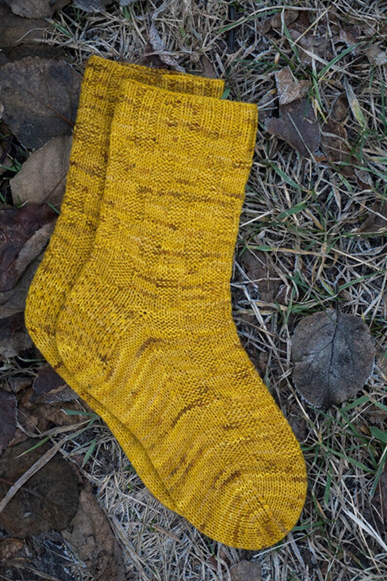 K&ouml;nnte beinhalten: Ein Paar gelbe Stricksocken mit einem strukturierten Muster. Die Socken liegen auf einem Bett aus trockenen Bl&auml;ttern und Gras.