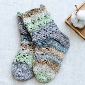 May include: A pair of hand-knitted socks in a multi-colored striped pattern with a lace design. The socks are made with a soft yarn in shades of green, brown, and gray.