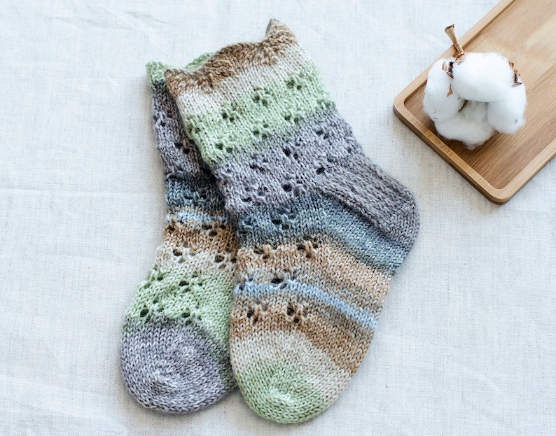 May include: A pair of hand-knitted socks in a multi-colored striped pattern with a lace design. The socks are made with a soft yarn in shades of green, brown, and gray.