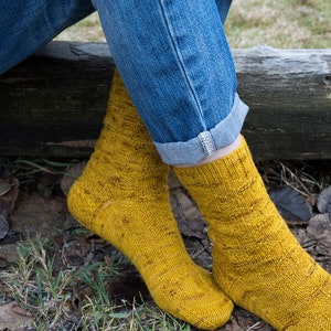 K&ouml;nnte beinhalten: Ein Paar gelbe Stricksocken mit einem strukturierten Muster. Die Socken werden von einer Person getragen, die auf einem Baumstamm sitzt.