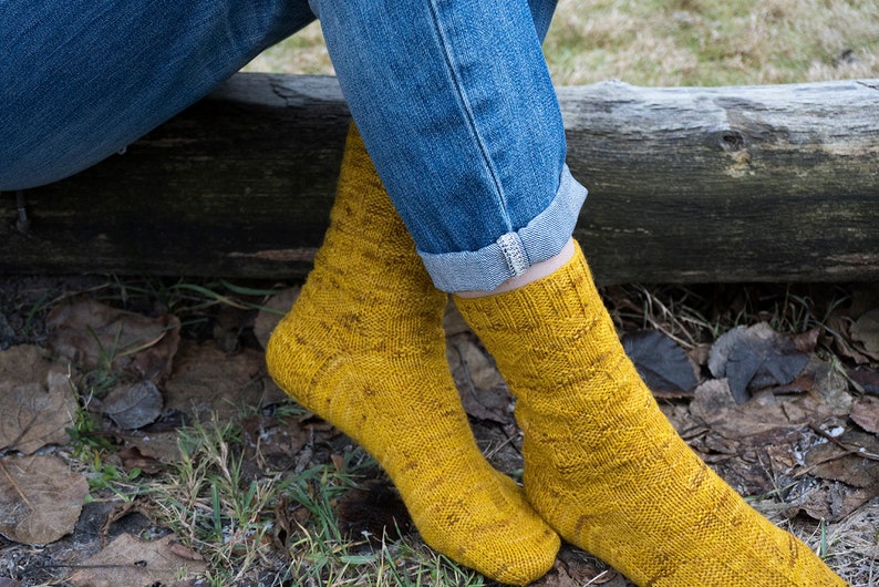 K&ouml;nnte beinhalten: Ein Paar gelbe Stricksocken mit einem strukturierten Muster. Die Socken werden von einer Person getragen, die auf einem Baumstamm sitzt.