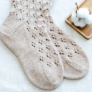 May include: A pair of beige knitted socks with a delicate lace pattern. The socks are lying on a white linen fabric background.