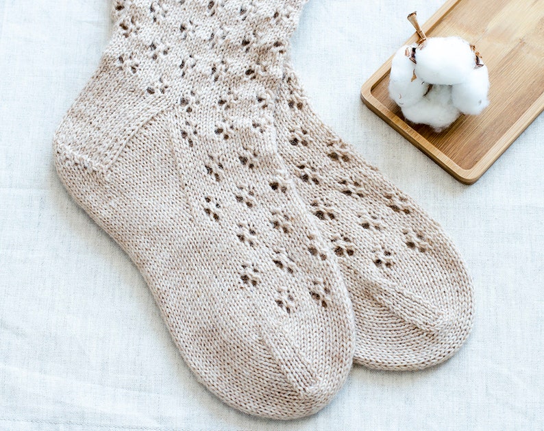 May include: A pair of beige knitted socks with a delicate lace pattern. The socks are lying on a white linen fabric background.