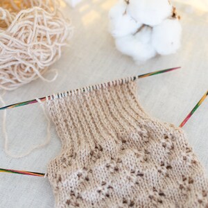 May include: A beige knitted garment with a lacy pattern is being worked on with two wooden knitting needles. The yarn is beige and is in a ball to the left of the garment.