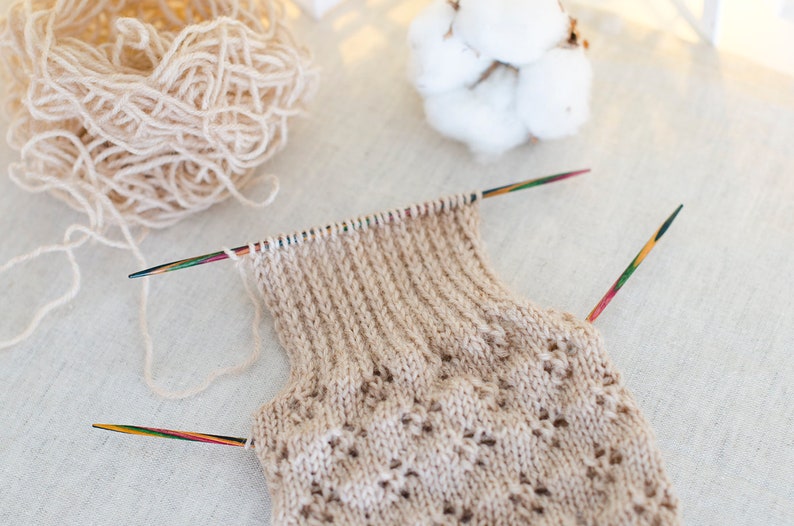 May include: A beige knitted garment with a lacy pattern is being worked on with two wooden knitting needles. The yarn is beige and is in a ball to the left of the garment.
