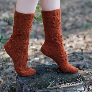 Op de afbeelding: Een paar bruine gebreide sokken met een bladmotief. De sokken worden gedragen aan de voeten van een persoon en staan op een boomstronk.