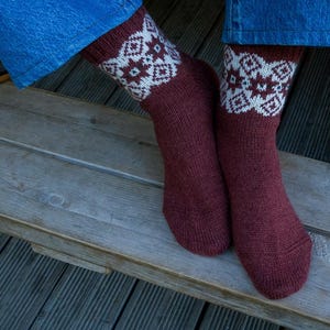 May include: A pair of knitted socks in a deep maroon colour featuring a white and maroon geometric snowflake pattern around the cuffs. The socks are worn with blue denim jeans, and the fabric shows a textured, hand-knit appearance throughout.