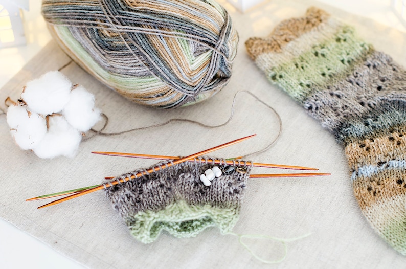 May include: A close-up of a knitting project with a ball of yarn, knitting needles, and a partially knitted sock. The yarn is a mix of brown, green, and gray colors. The sock is also a mix of brown, green, and gray colors. The knitting needles are wooden and are resting on a white surface.