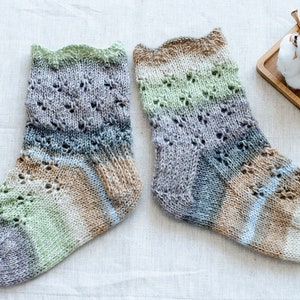 May include: A pair of hand-knitted socks in a multi-colored yarn with a lace pattern. The socks are made with a variety of shades of green, brown, blue, and gray.