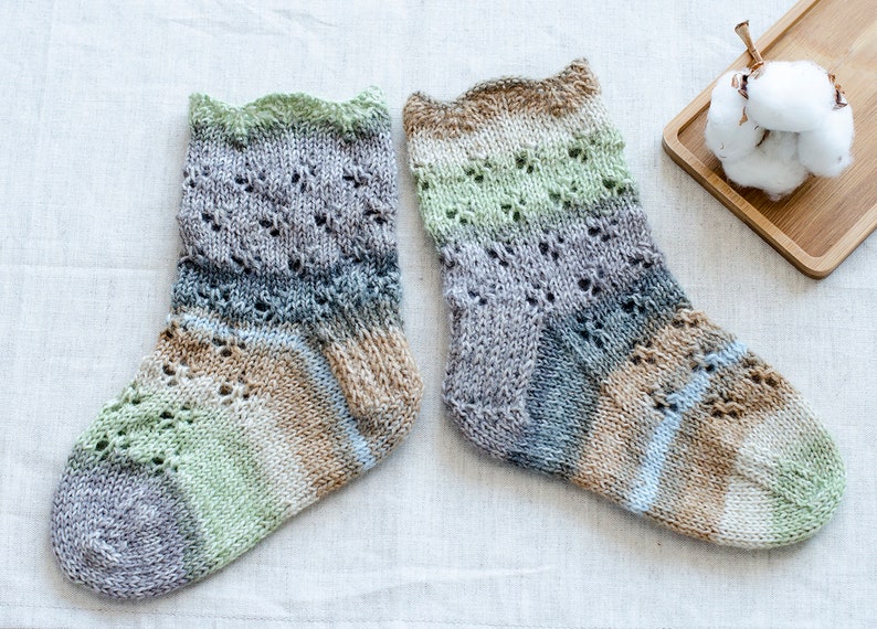 May include: A pair of hand-knitted socks in a multi-colored yarn with a lace pattern. The socks are made with a variety of shades of green, brown, blue, and gray.