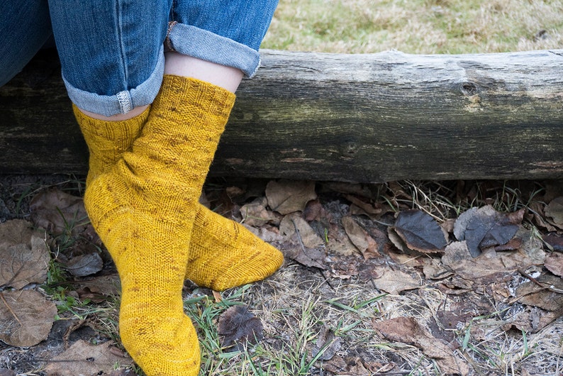 K&ouml;nnte beinhalten: Ein Paar gelbe Stricksocken mit einem strukturierten Muster. Die Socken werden von einer Person getragen, die auf einem Baumstamm mit heruntergefallenen Bl&auml;ttern sitzt.