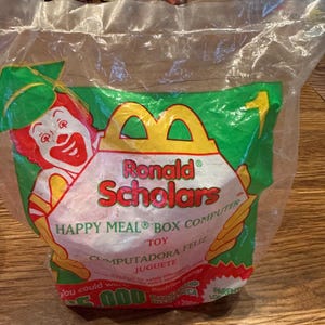 May include: Sealed Happy Meal toy bag featuring Ronald McDonald in a graduation cap. The bag is green and yellow with the words "Ronald Scholars" and "Happy Meal Box Computer" printed on it. The bag is made of clear plastic.