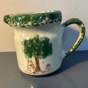 May include: A small, white ceramic pitcher with a green rim and handle. The pitcher features a hand-painted design of a tree with pink fruit, a ladder, and a basket of purple flowers. The handle and rim have a green speckled design.
