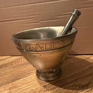Puede incluir: Un juego de mortero y mano de bronce. El mortero tiene la palabra "GALEN" grabada alrededor del borde. La mano descansa dentro del mortero. El juego está sobre una superficie de madera.