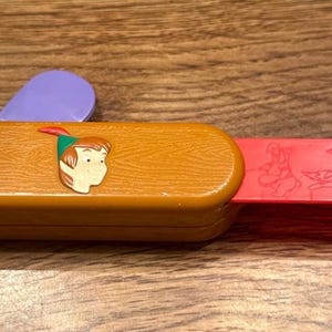 May include: A vintage Peter Pan toy featuring a brown rectangular body with a Peter Pan character image, green handles, a purple piece, and a red sliding piece with character silhouettes. The toy is made of plastic.