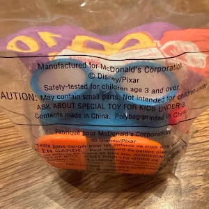 Może przedstawiać: Zgrzana plastikowa torba zawierająca niebieską zabawkę z pomarańczowymi akcentami. Opakowanie zawiera tekst od Disney/Pixar i McDonald's Corporation. Zabawka jest testowana pod kątem bezpieczeństwa dla dzieci w wieku od 3 lat.