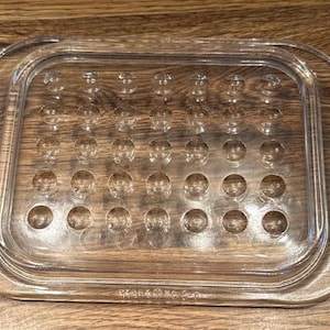 May include: A rectangular clear glass butter dish lid featuring a grid of thirty-five circular indentations on the top surface. The lid has a smooth, raised border and a flat, wide rim, with embossed text visible along the bottom edge of the glass.