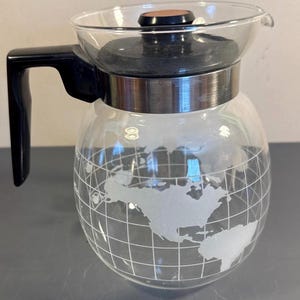 May include: A clear glass coffee carafe featuring a frosted white etched map of the world with a grid pattern. The carafe includes a black plastic handle, a stainless steel band around the neck, and a black lid with a small brown circular accent.