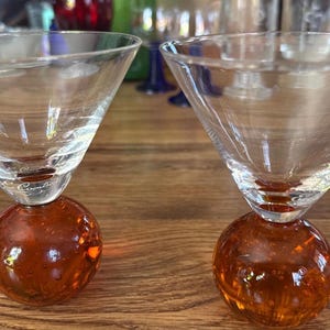 May include: Two clear martini glasses with a conical bowl and a round, amber-colored base. The glasses are on a wooden surface, showcasing their unique design and color contrast. The stem of the glass has a brand name.