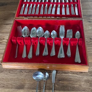 Puede incluir: Un estuche de madera para cubiertos con interior de terciopelo rojo que contiene un juego de cuchillos, tenedores y cucharas plateados. Tres utensilios adicionales, incluyendo un cucharón, una cuchara pequeña y un cuchillo de mantequilla, están colocados fuera de la caja.