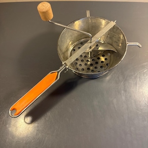 May include: A vintage food mill with a perforated metal bowl, a rotating blade, and an orange handle. The mill has a wooden knob and a metal handle for turning. The bowl is approximately 8 inches in diameter.