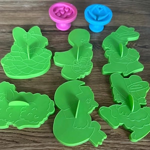 Puede incluir: Un conjunto de cortadores de galletas de plástico verde con temática de Pascua, que incluyen un conejito, un pollito y un cordero. También hay dos sellos coloridos, uno rosa con una huella de pata y otro azul con una flor. Dispuestos sobre una superficie de madera.