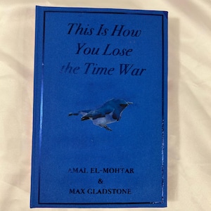 Può includere: Un libro blu con copertina rigida intitolato "This Is How You Lose the Time War". La copertina presenta un'illustrazione di un uccello blu e i nomi degli autori, Amal El-Mohtar & Max Gladstone. Il libro è su uno sfondo bianco.