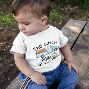 Peut inclure: Un tout-petit portant un t-shirt blanc avec un camion bleu et des citrouilles dessus. Le t-shirt dit "The Cutest Pumpkin in the Patch". Le tout-petit est assis sur un banc en bois.