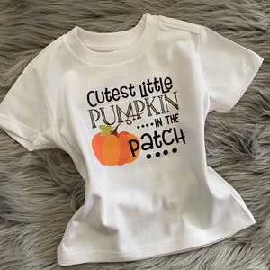 May include: White short-sleeved t-shirt with a black graphic that says "Cutest little pumpkin... in the patch" with a small orange pumpkin illustration.