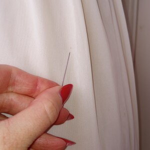 May include: A close-up of a white fabric with a small pin inserted into it. The pin is held by a hand with red nail polish.