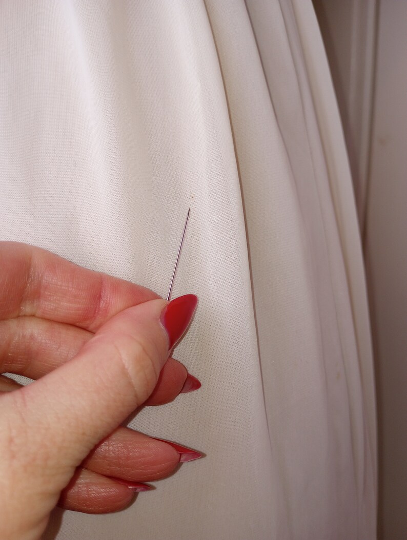 May include: A close-up of a white fabric with a small pin inserted into it. The pin is held by a hand with red nail polish.