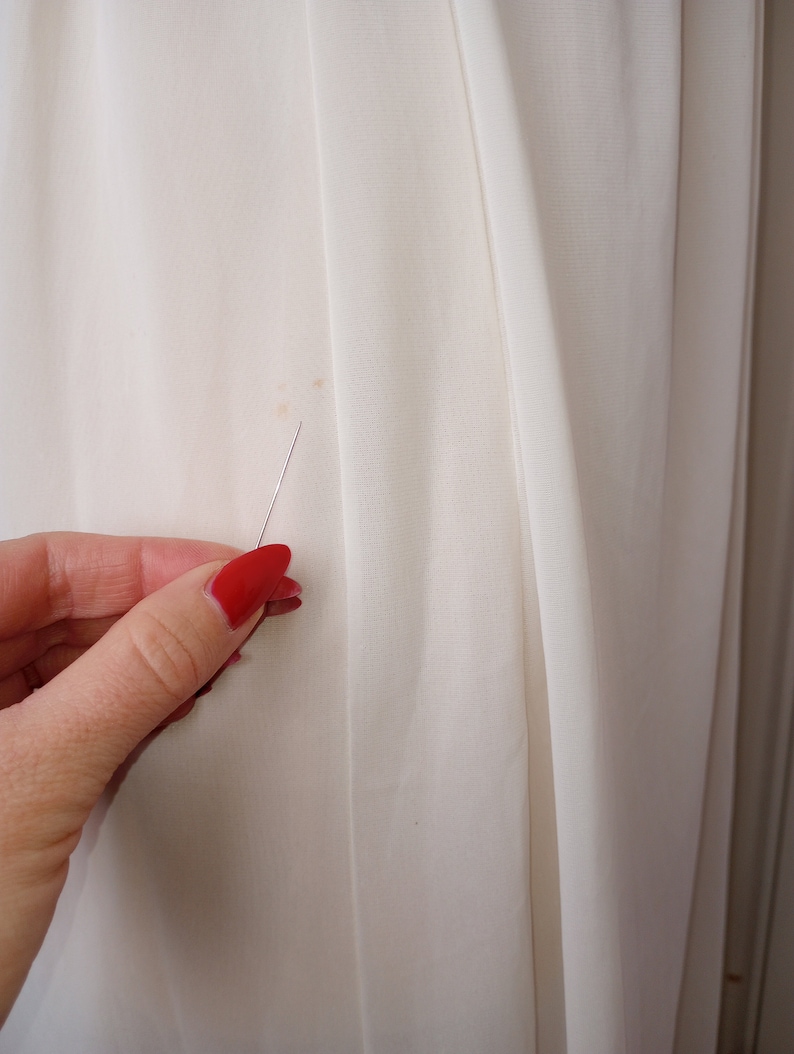 May include: Close-up of a white fabric with a small brown stain. A hand with red nail polish is holding a pin near the stain.