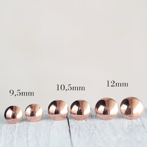 May include: Five rose gold dome stud earrings in different sizes: 9.5mm, 10.5mm, 12mm. The earrings are arranged in a row from smallest to largest on a white wooden surface.