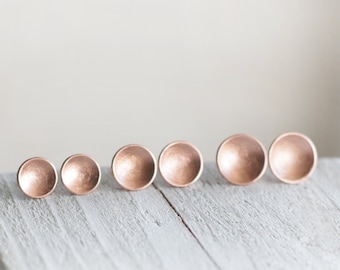 Puede incluir: Cinco pendientes redondos en forma de disco de color oro rosa, de diferentes tamaños, dispuestos en fila sobre una superficie de madera blanca.