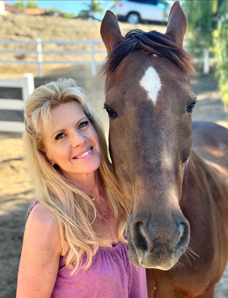 May include: A woman with blonde hair smiles next to a brown horse with a white diamond-shaped marking on its forehead. The woman wears a pink top. The background includes a white fence and a vehicle.